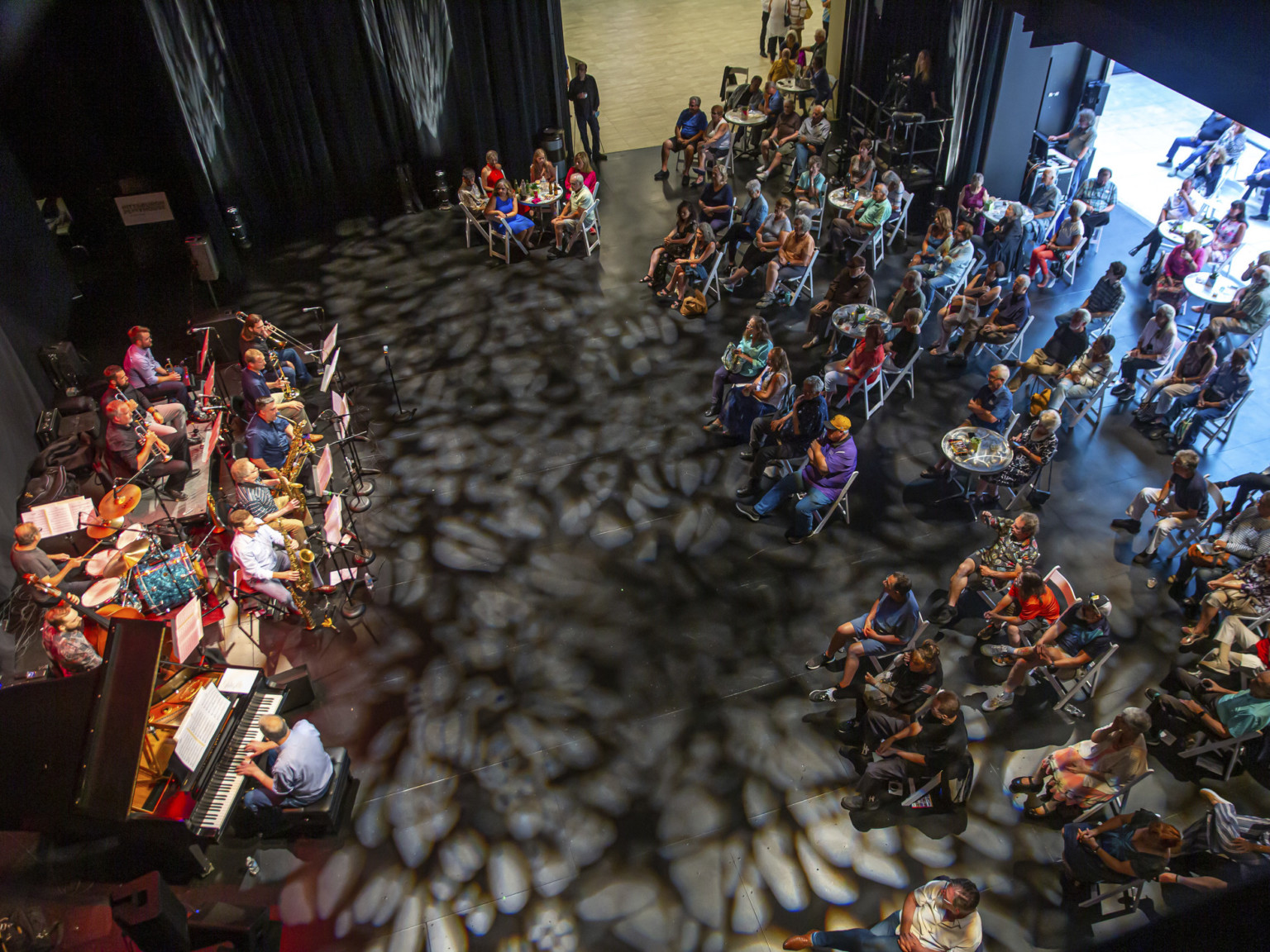 Audience sits in scattered folding chairs facing musicians while texture white light is projected onto the black floor