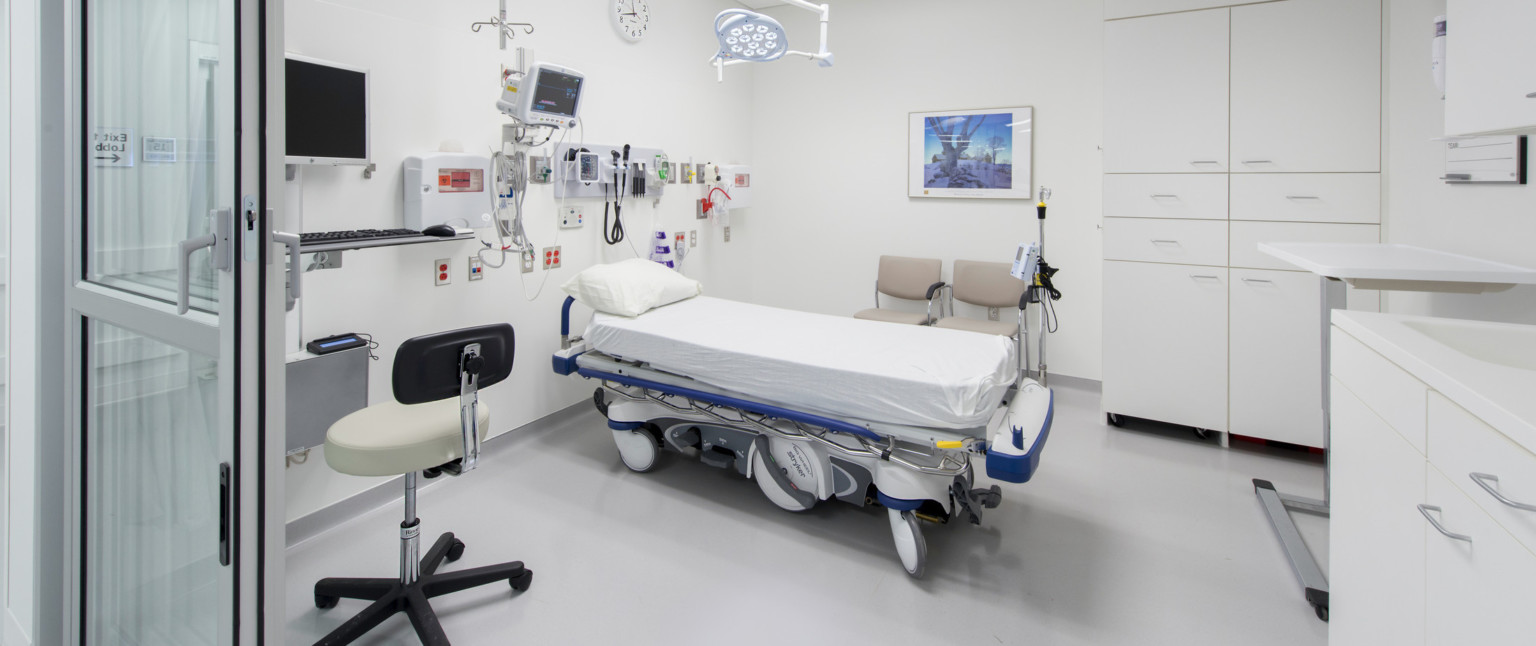 Hospital bed in white room with equipment over head. Two beige chairs behind bed next to 2 large cupboards. Desk on left wall