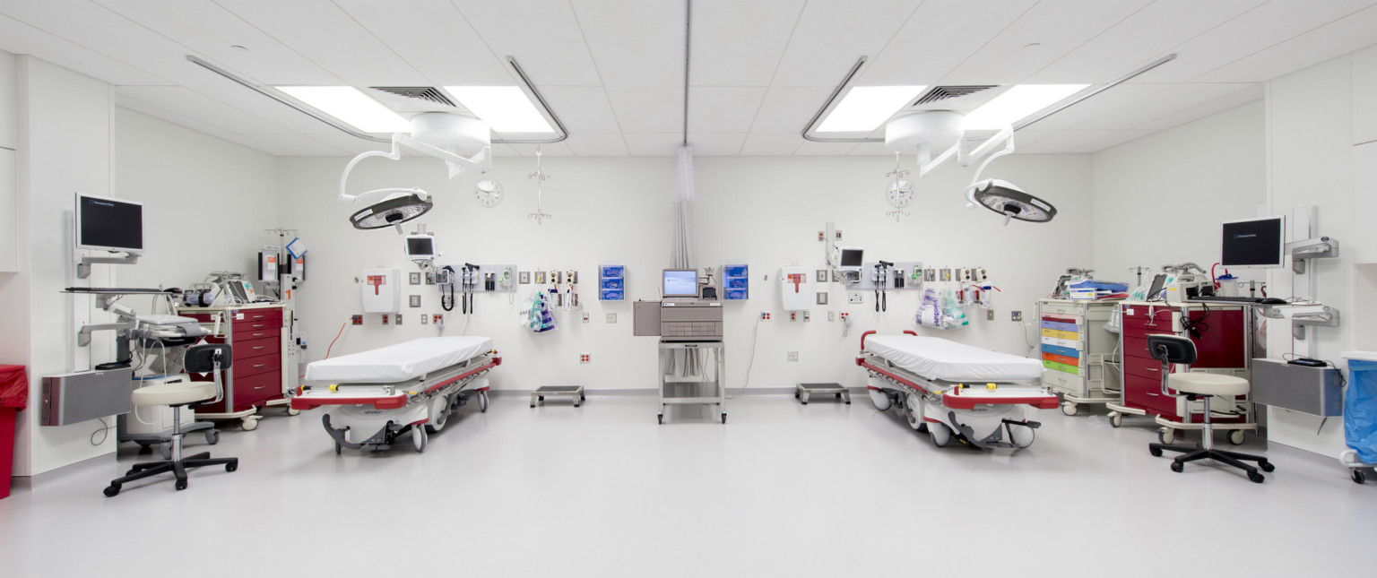 White room with 2 hospital beds with large circular whites above. Machine on rolling table center. Medical carts on each side