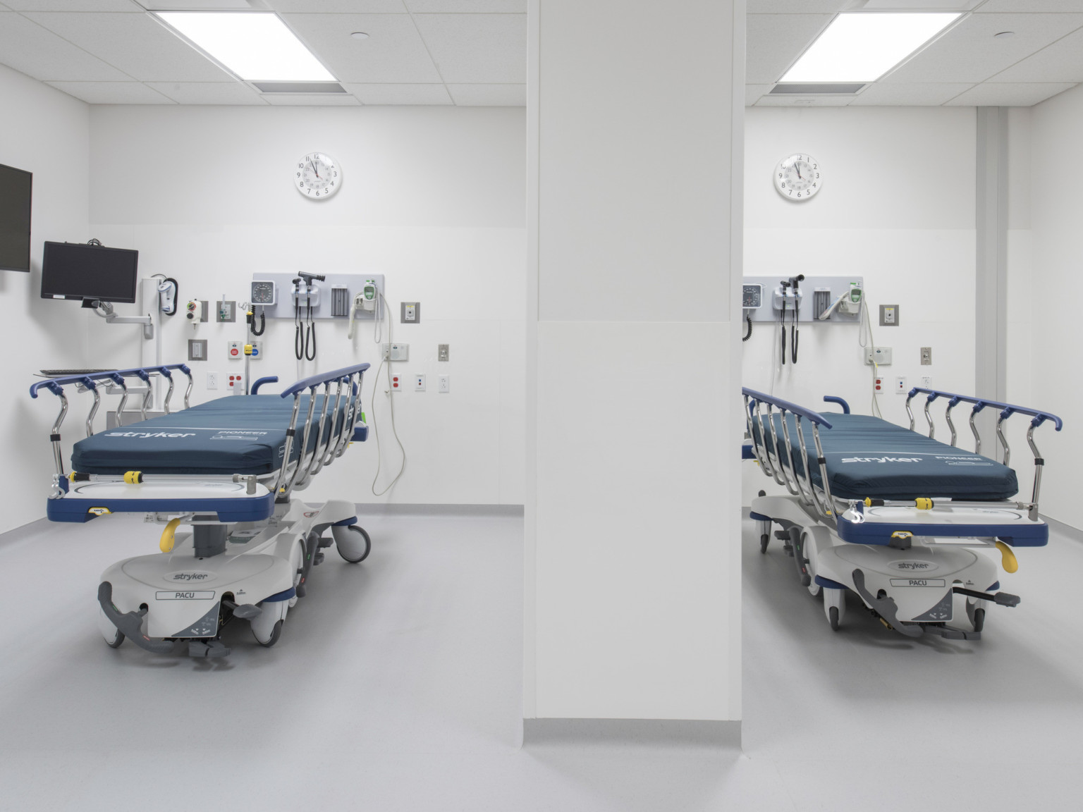 Two hospital beds reclined in a white room with a pillar. Equipment of vitals hangs on wall above each, with a clock on top
