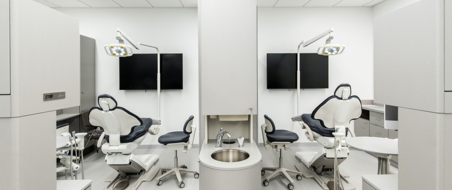 Two dental chairs with seat in between facing a screen, with movable light hanging above. Faucet sits in between the chairs