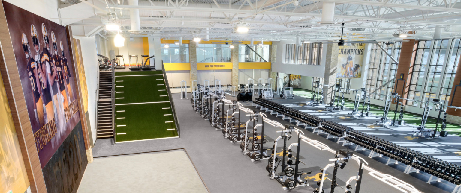 Double height training facility with rows of equipment. Left, a Wyoming Football mural on wall beside stairs and turf slope