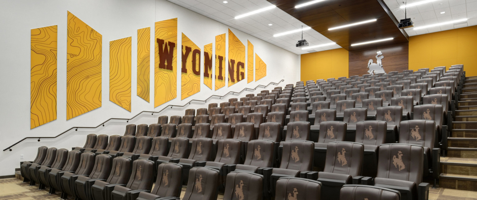 Brown theater style stadium seats with logos in white room with yellow mural reading Wyoming on left wall. Yellow back wall