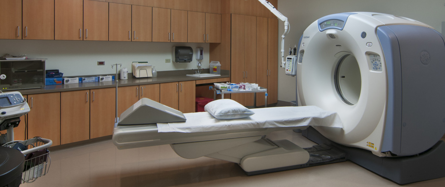 MRI machine in center of white room with wood cabinets right of shelves above and below counter space with sink