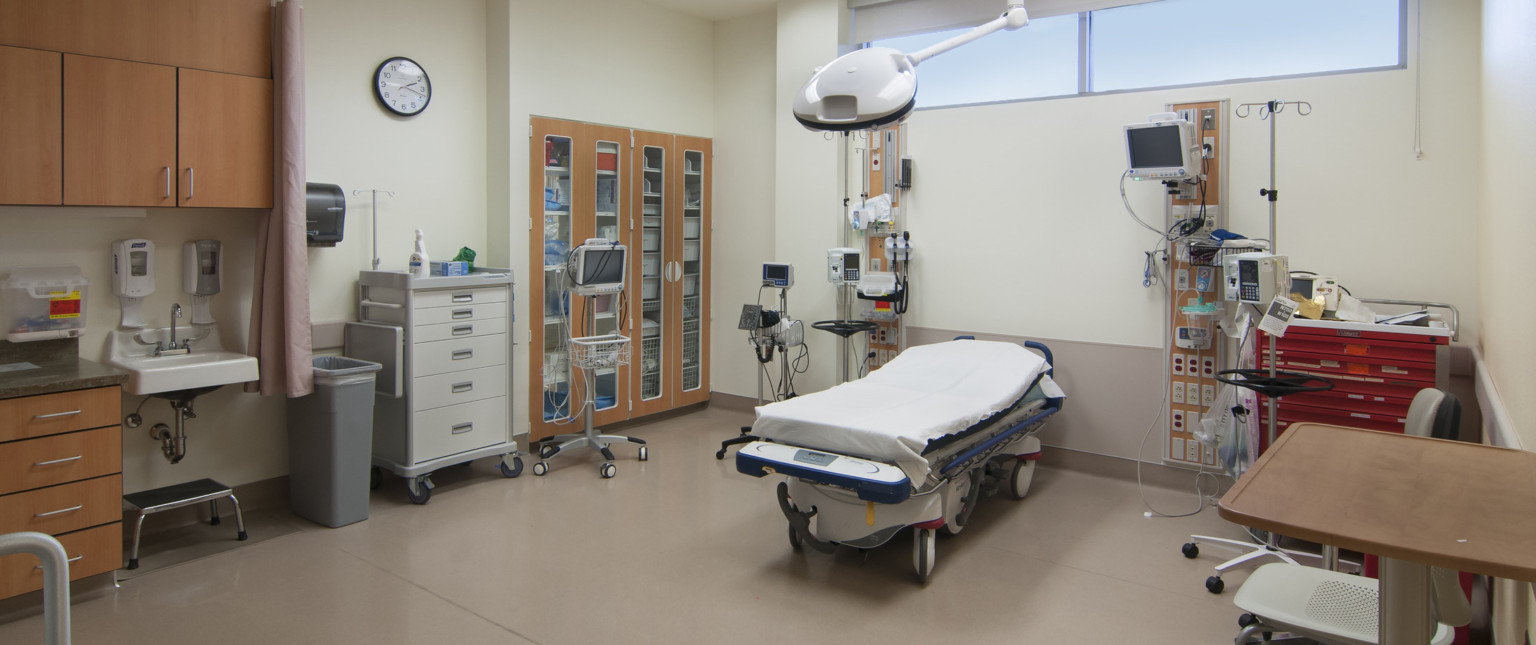 Hospital bed center equipment on wall on both sides. Built in cupboard with wood doors on left wall next to medical cart
