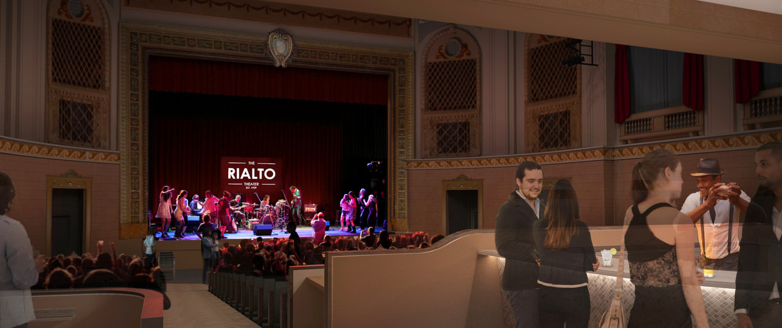 People stand at the bar in front right of image while others sit watching performance on stage of historic theater