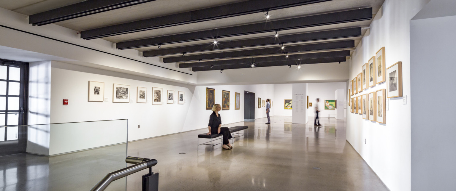 White gallery hall with paintings on the walls and benches along the center. Black beams cross the room's width with lighting