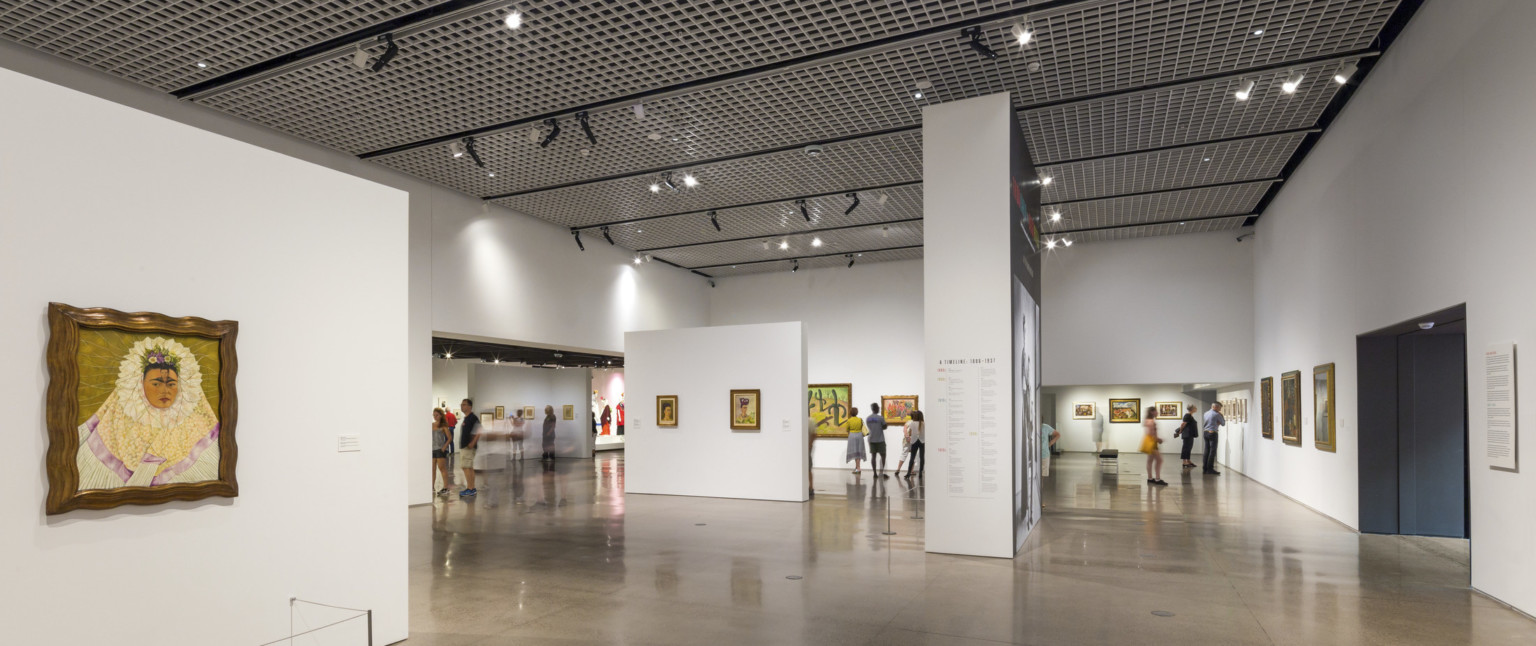 A steel grid with light fixtures hanging off spans the ceiling above a white gallery with paintings on the walls