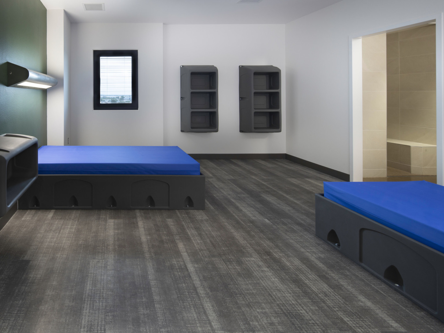 Dark flooring in white room with green accent wall. 2 blue beds on grey bases with light above. Grey shelves on far wall