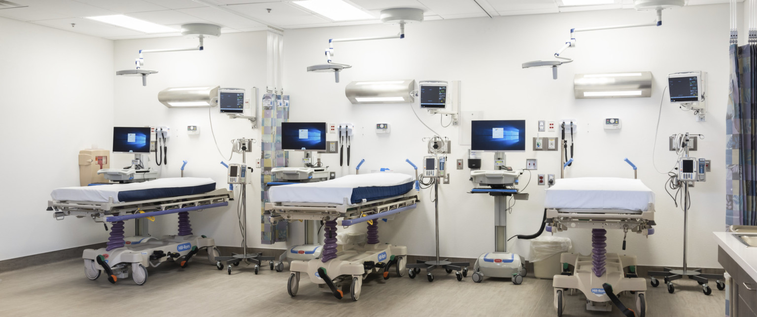White room with hospital beds, light above, equipment right, and portable computers left. Patterned curtain hangs for privacy