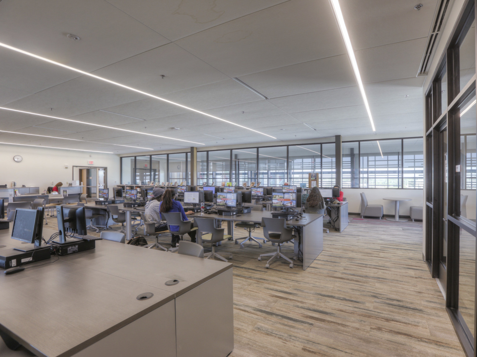 Computer lab space with lines of recessed lighting over wood floors. Glass wall to right, windows line upper back wall