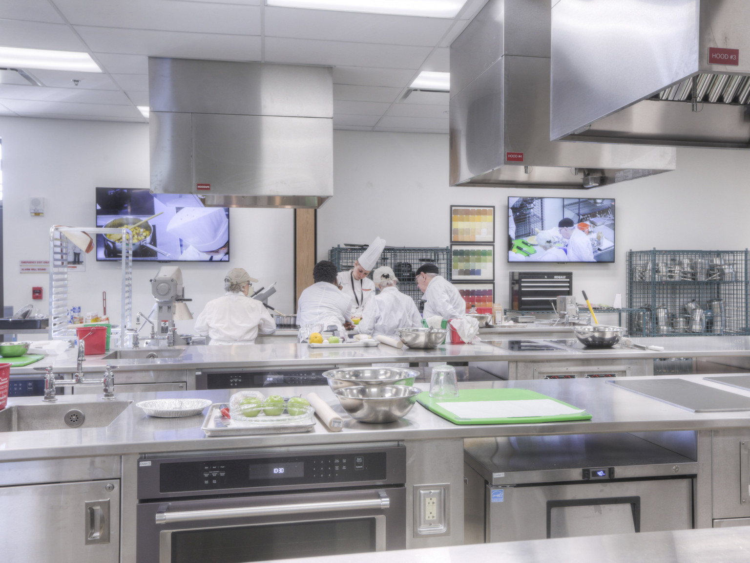 Teaching kitchen with stainless steel counters and appliances facing front with screen showing close up view of demonstration