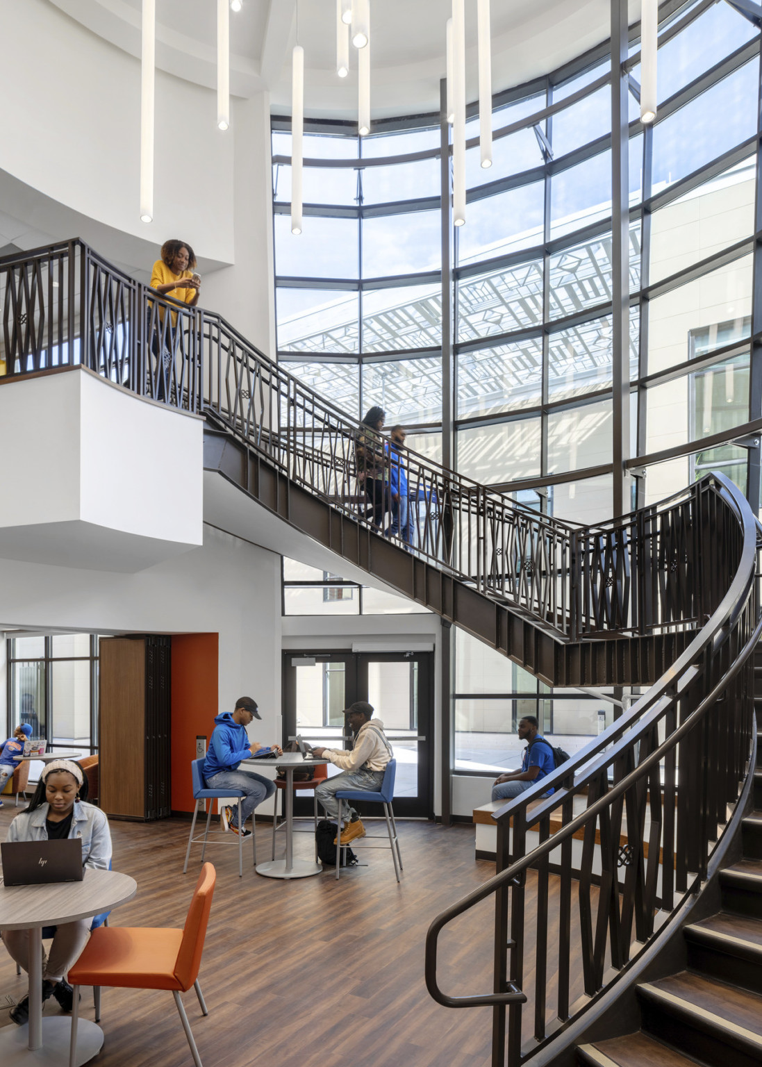 Curved staircase along double height windows. Above, cylindrical white pendant lights, seating area at the base of the stairs