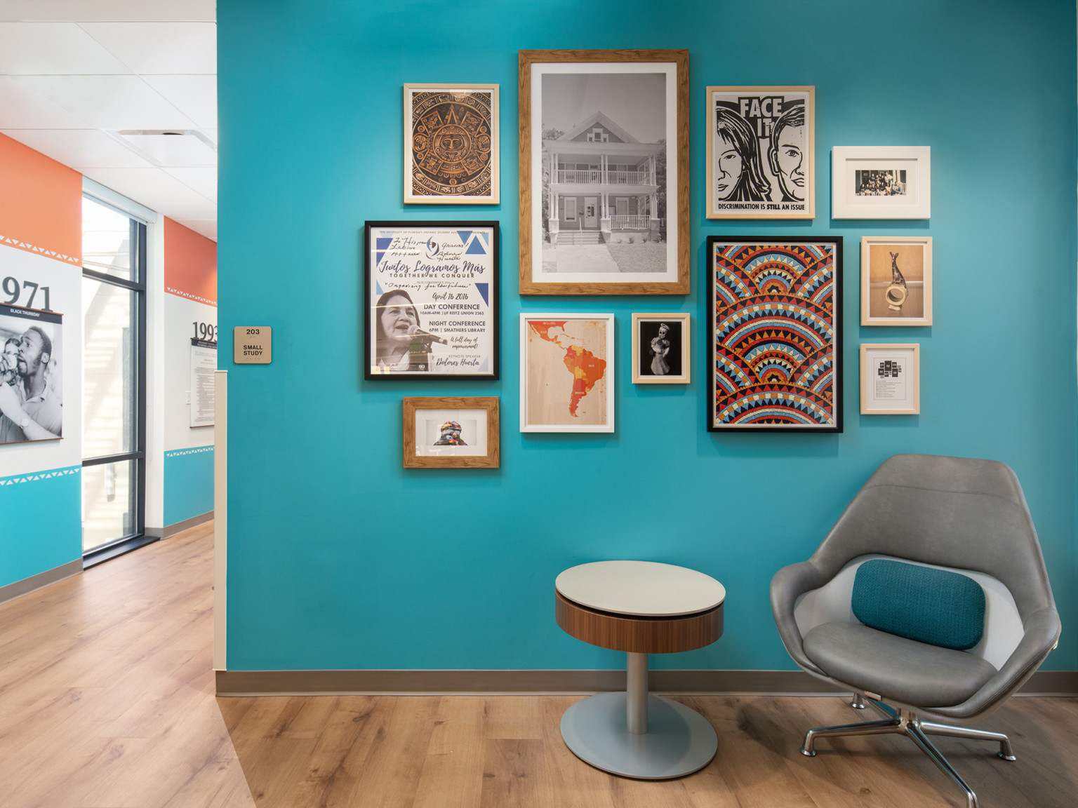 Teal gallery wall with framed art behind grey chair and round side table. Hallway connects at left with multicolored wall