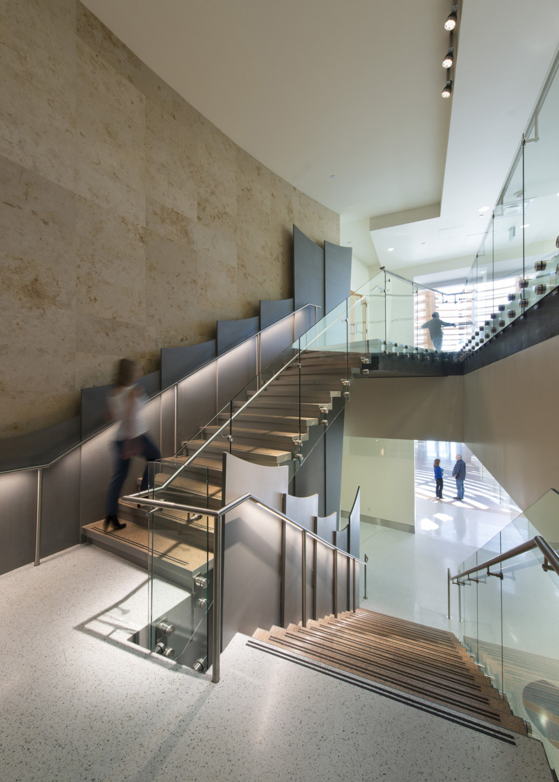 A stairwell between 2 white hallways with natural lighting. Curved grey panels line the left wall past the glass rail