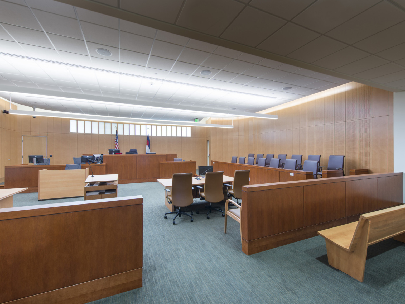A courtroom with dark teal carpeting and wood paneled walls. A thin window sits above the judge's desk with 2 flags between