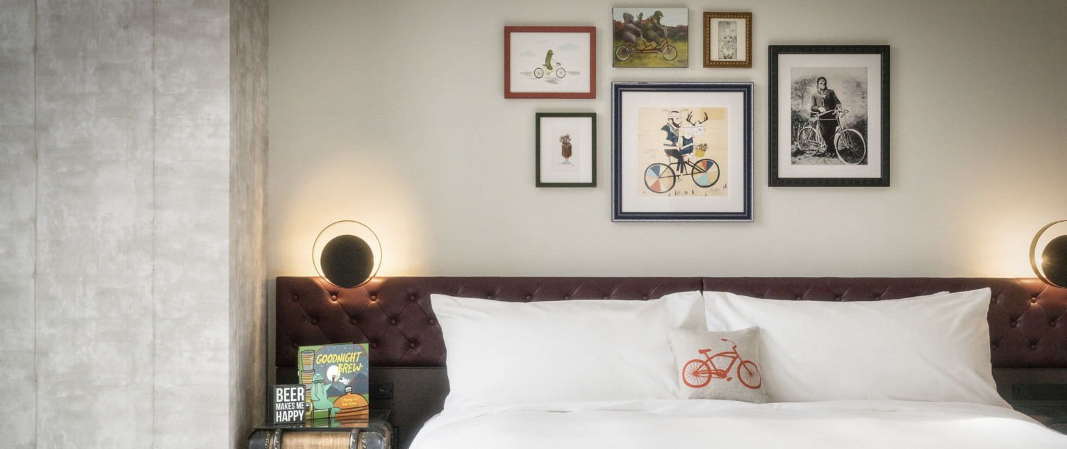 White bed with brown tufted headboard and round lamp. Bike themed art framed on wall and throw pillow on bed