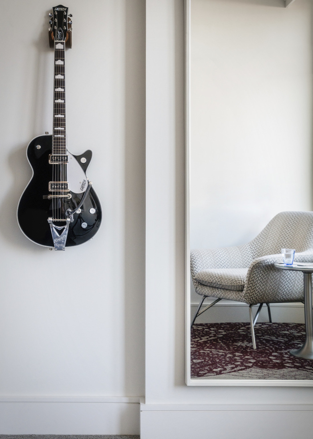 White room with black and white guitar hanging on wall next to mirror reflecting white textural upholstered chair on red rug