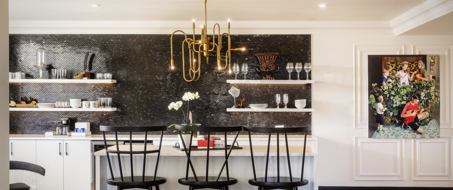 Closeup of kitchen. Black tile accent over white counter. A gold organically curved chandelier hangs over a white island