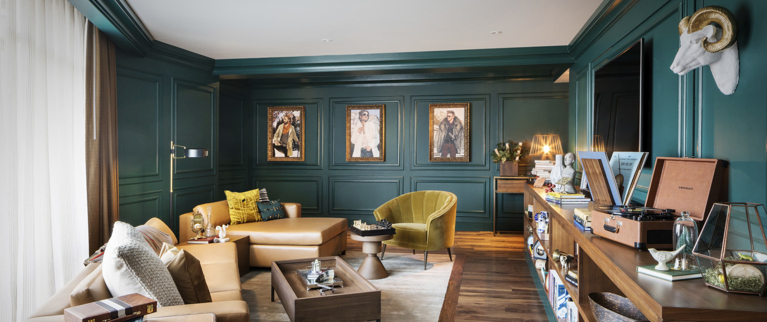 Sitting room with dark green molded walls. Light brown leather couch and yellow velvet chair with wood coffee table and shelf