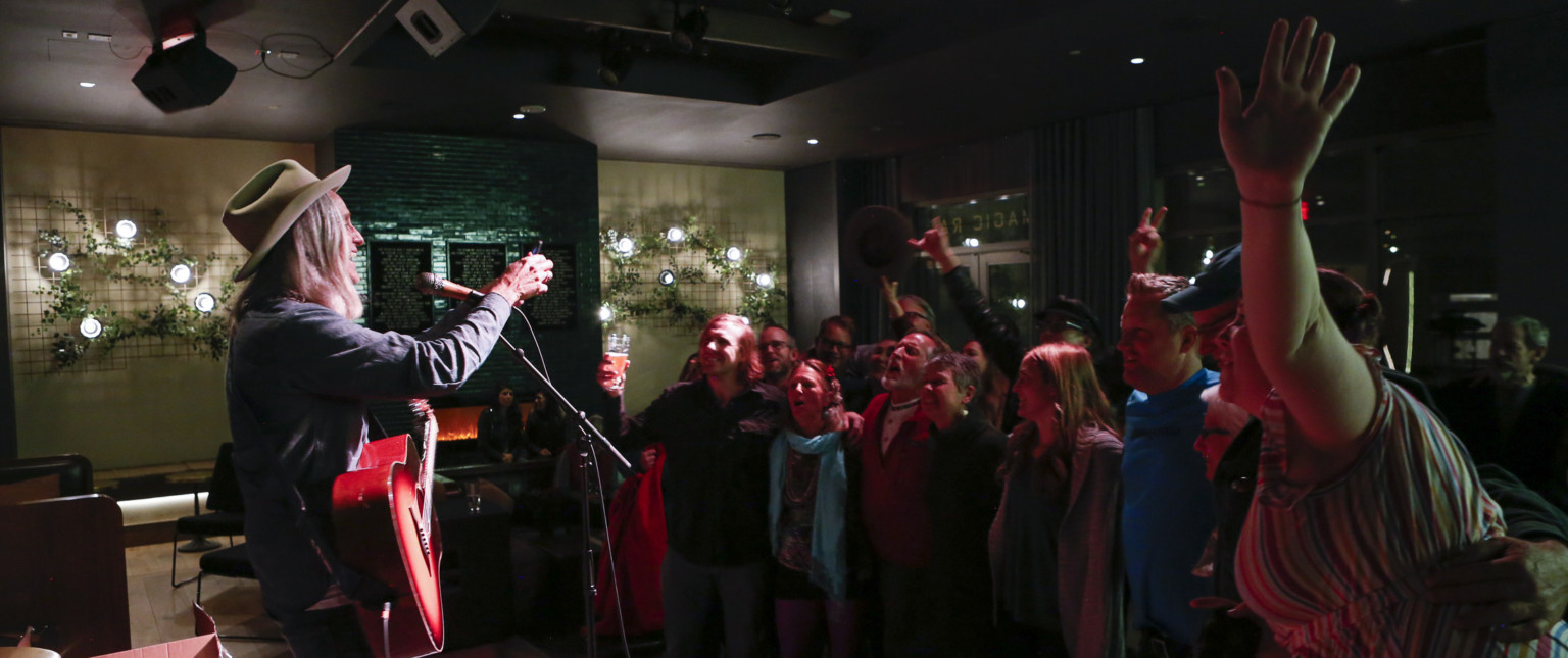 Man performs for crowd in room with green fireplace illuminated by grid with greenery and lighting on white walls by fire