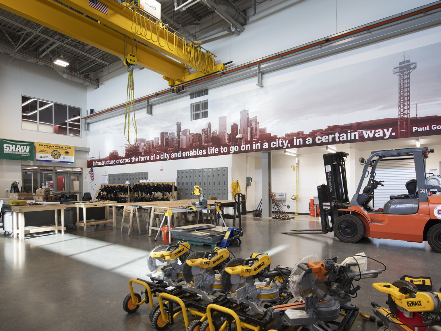 Double height mechanical lab with forklift to back right. Mural above with quote on infrastructure. Rows of wood tables, left