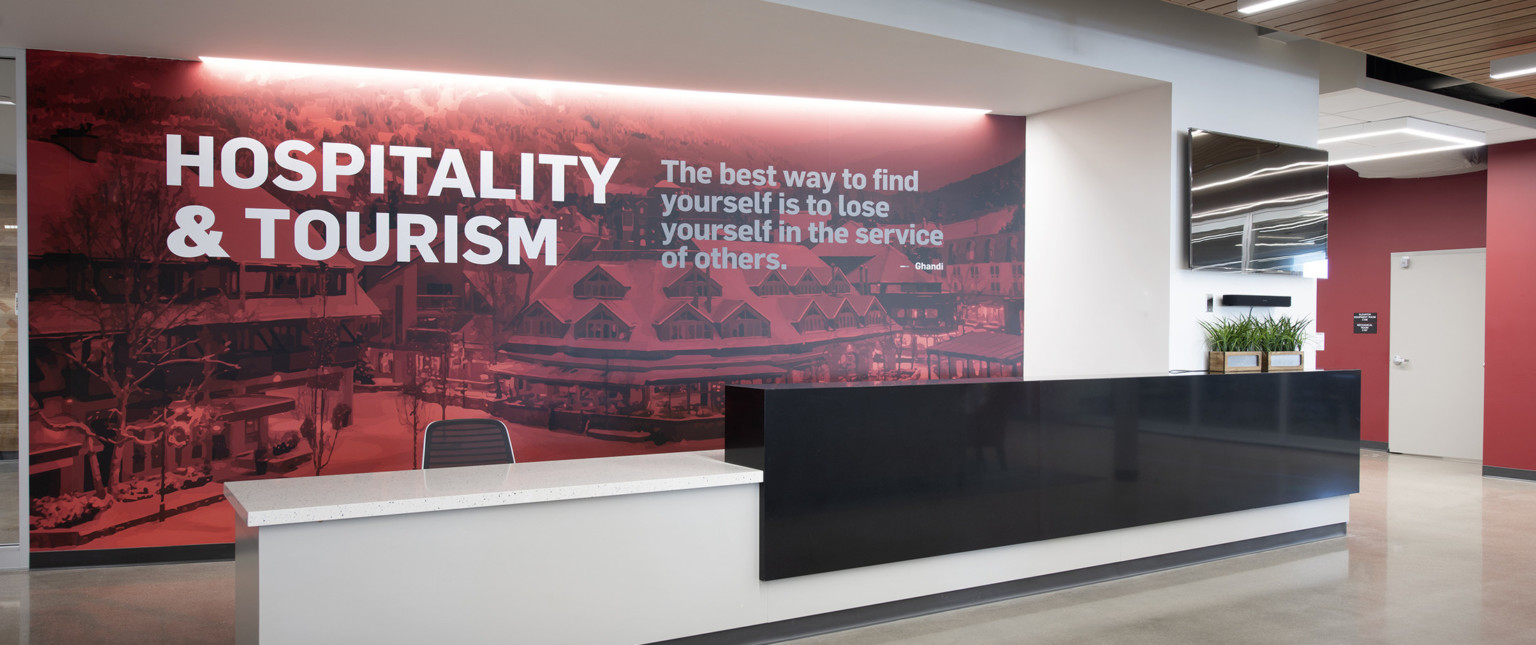 White desk with black partition in front of recessed wall mural with writing: Hospitality and Tourism, and quote from Ghandi
