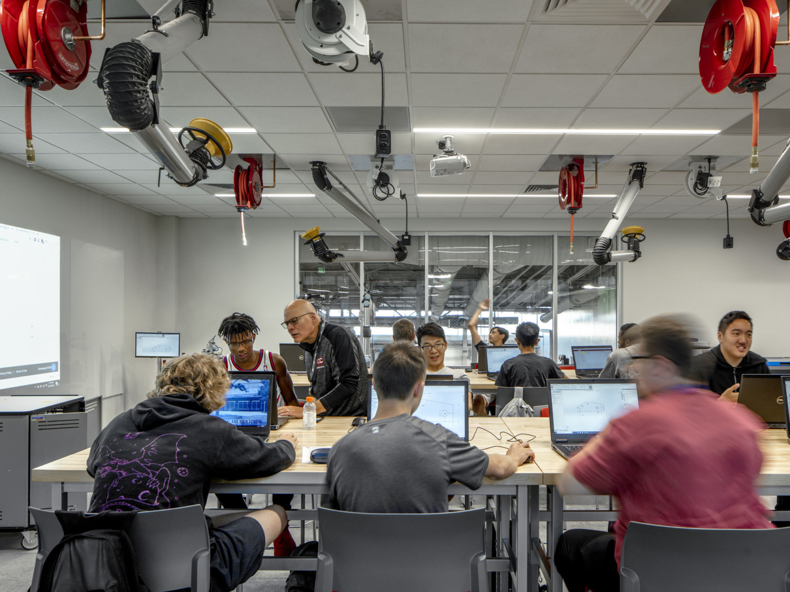 Classroom with students on computers at rows of wood tables. Equipment and power cords hang from the ceiling of white room