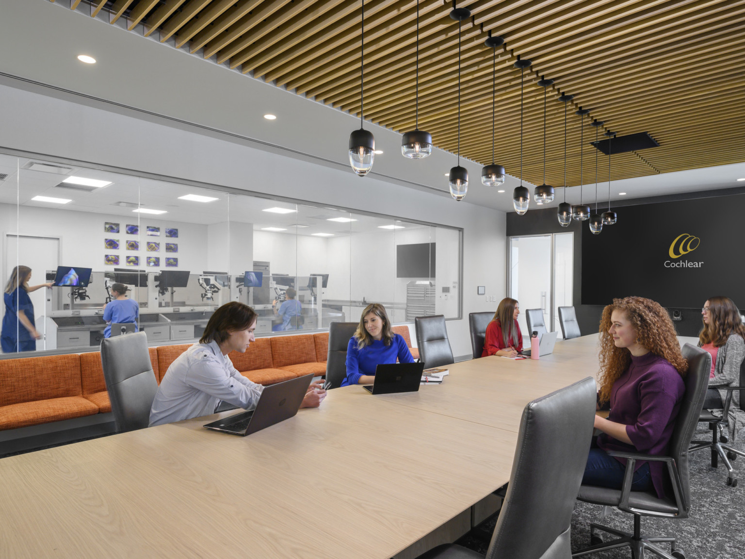 Conference room with wood slat ceiling over long table with pendant lights down center. Black back wall with large screen