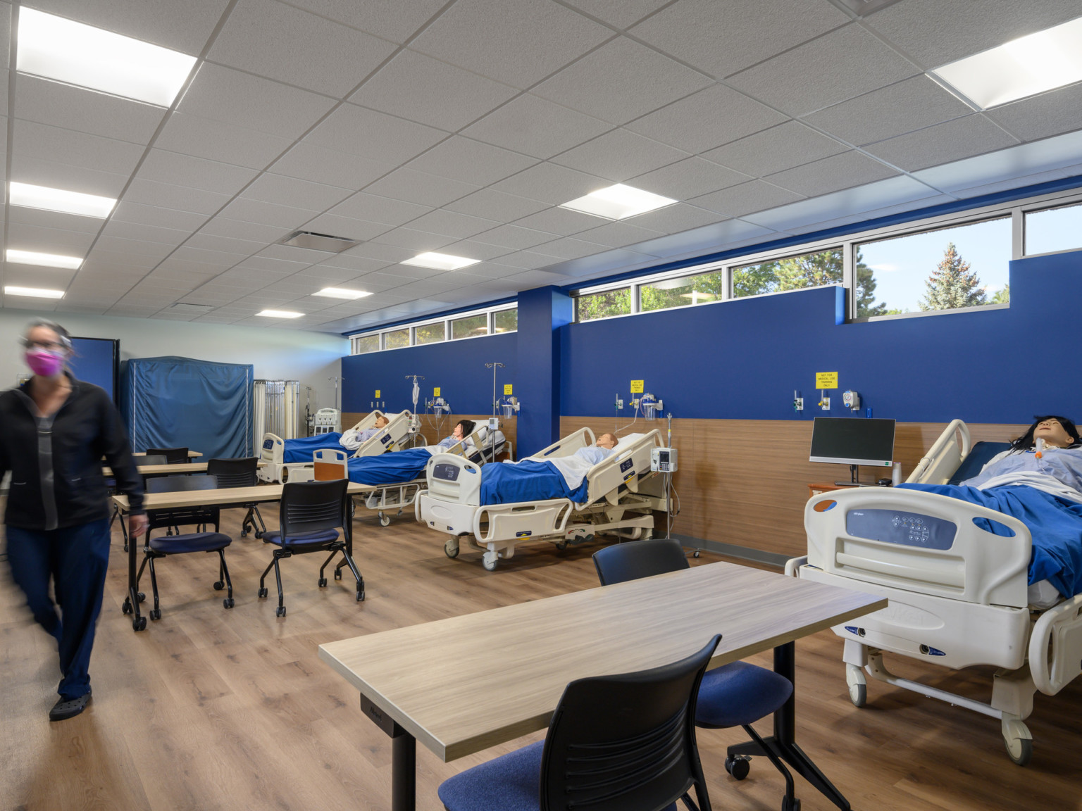 Hospital beds with dummies along wood wall with window lined blue accent upper half. Tables in front of beds on wood floor