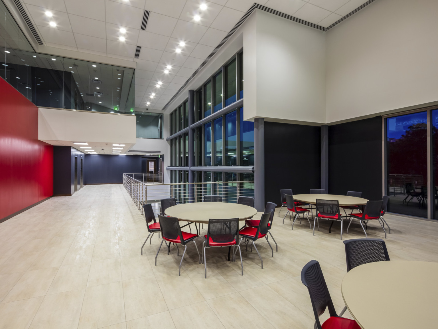 Double height window lined room with round tables and chairs. Red wall left and overhang with floor to ceiling windows above