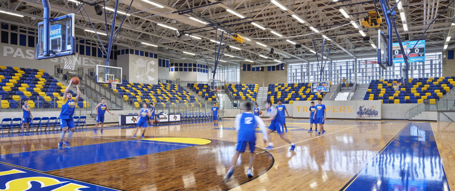 Wood basketball court surrounded by blue and yellow bleachers. Murals with Troopers mascot. Exposed structural beams above