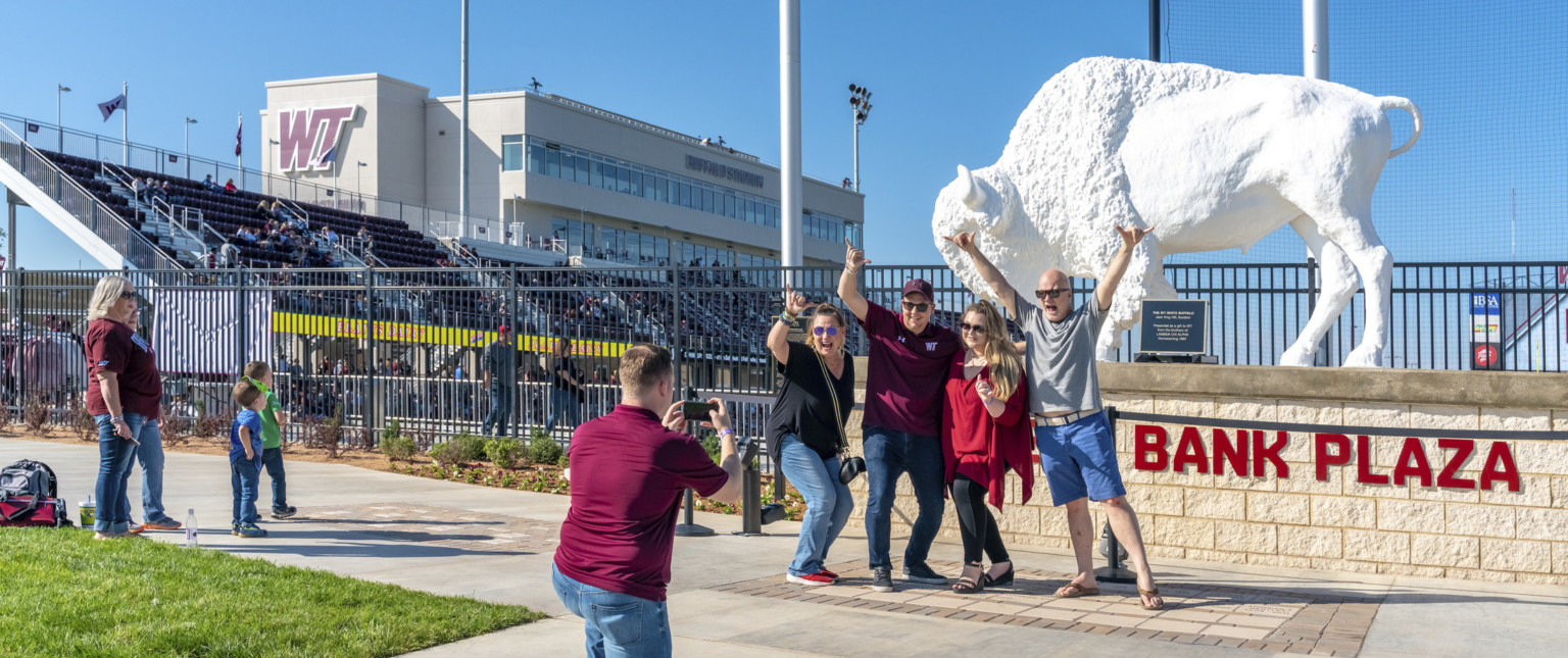 White buffalo sculpture on stone platform outside of black metal stadium fence. Behind, bleachers and box seats are visible