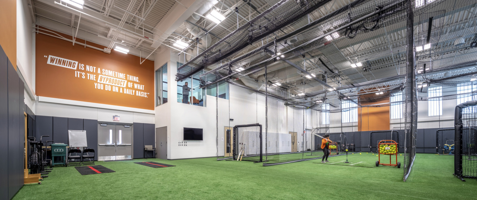 Indoor training space with turf flooring and netted batting cages to the right. Windows to 2nd floor viewing room by mural