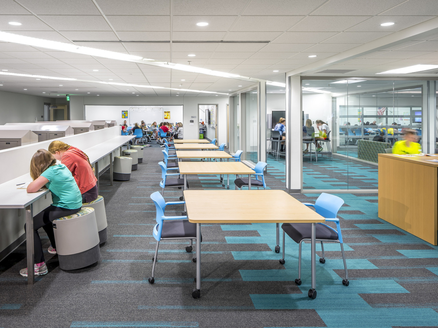 Long white room with white counter and stools, left. Tables with blue chairs, right. Glass walled learning spaces far right