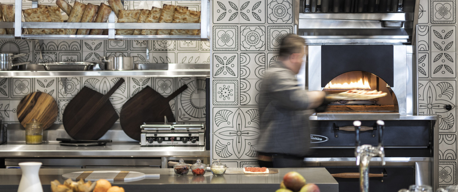 Pizza being placed into oven in kitchen with black and white floral tiles. Stainless steel counters and shelving with bread
