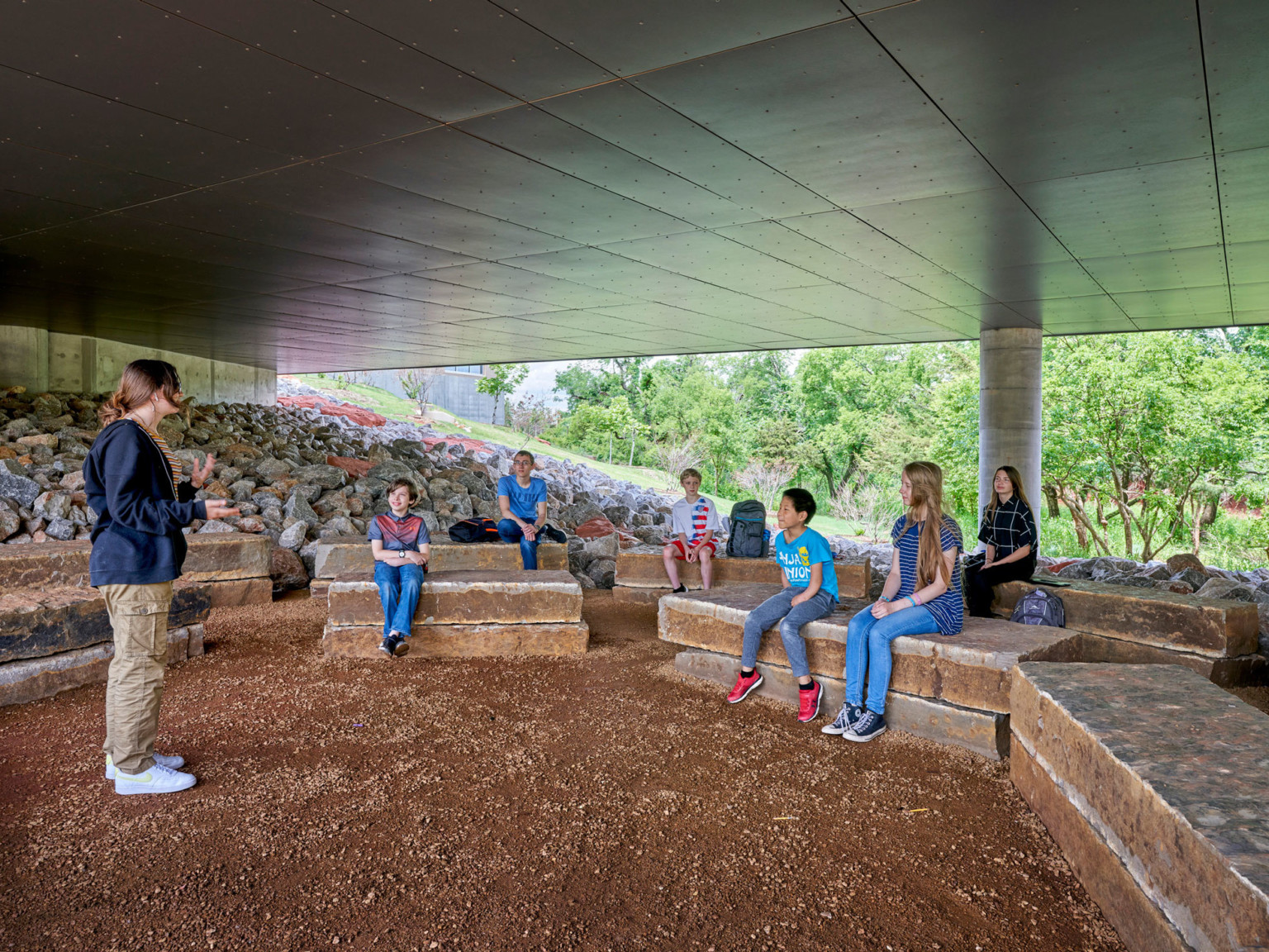 Outdoor learning space with rock benches arranged in semi circle, covered by hallway and open at sides