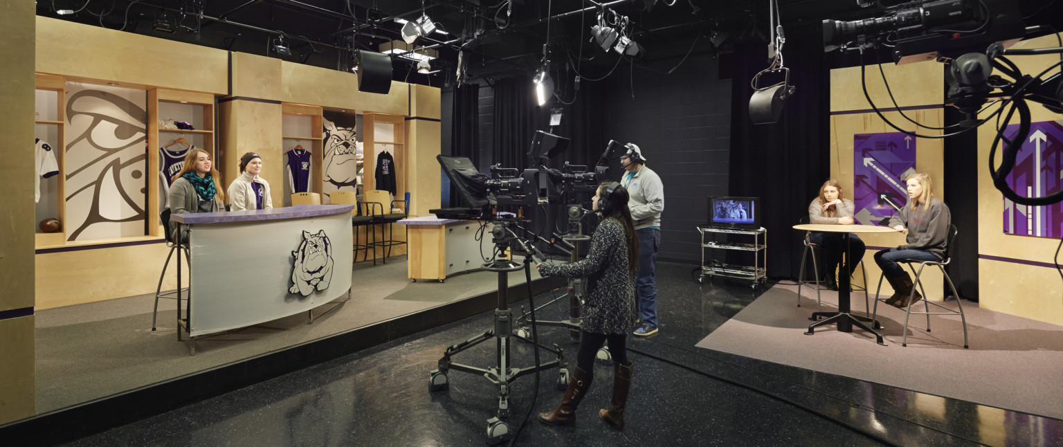 A newsroom with wood wall and desk, accented with purple and images of the bulldog mascot. 2 cameras face the stage