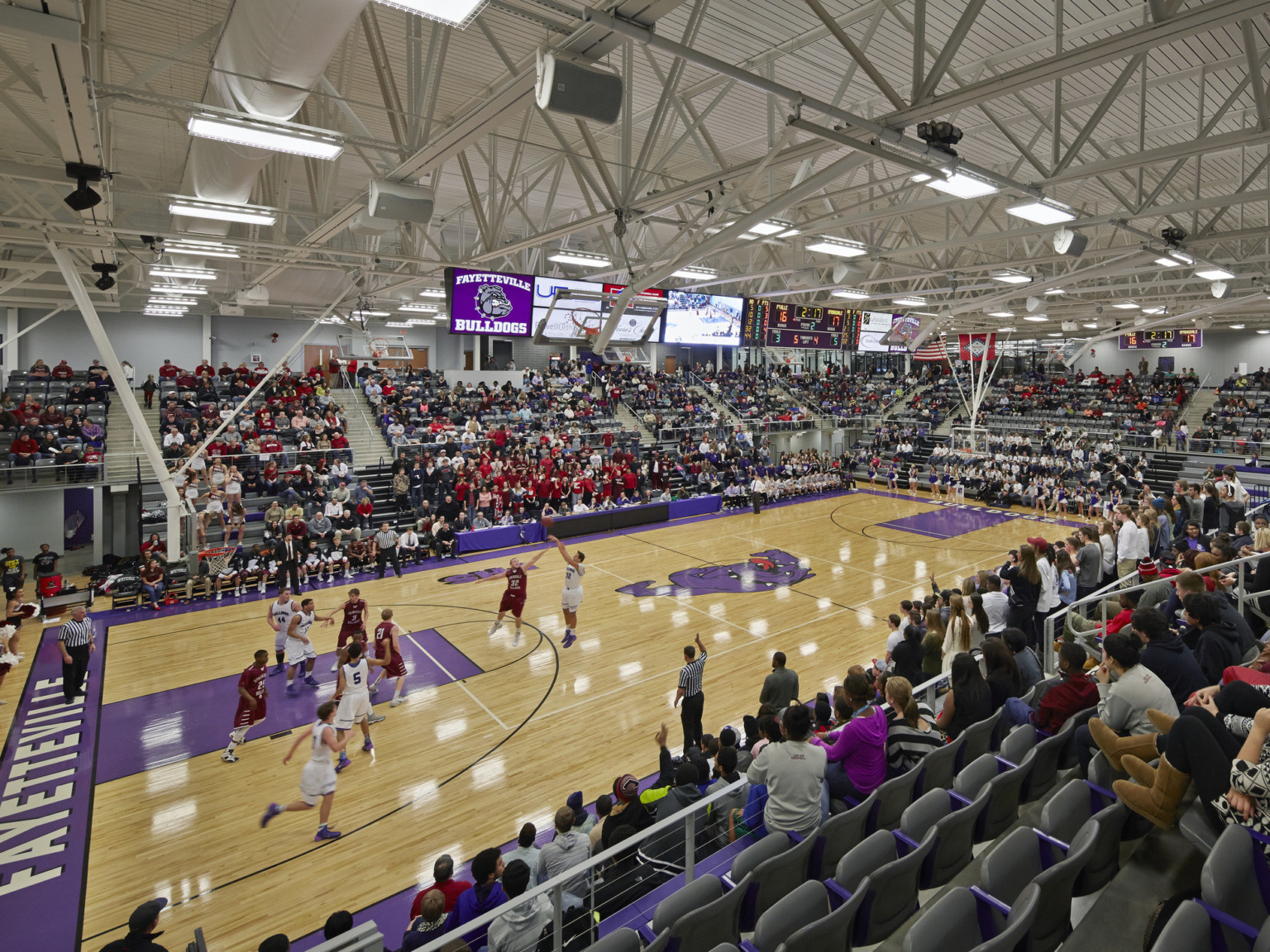Double height gymnasium in use during a Fayetteville Bulldogs basketball game. Bleachers surround wood floor basketball court