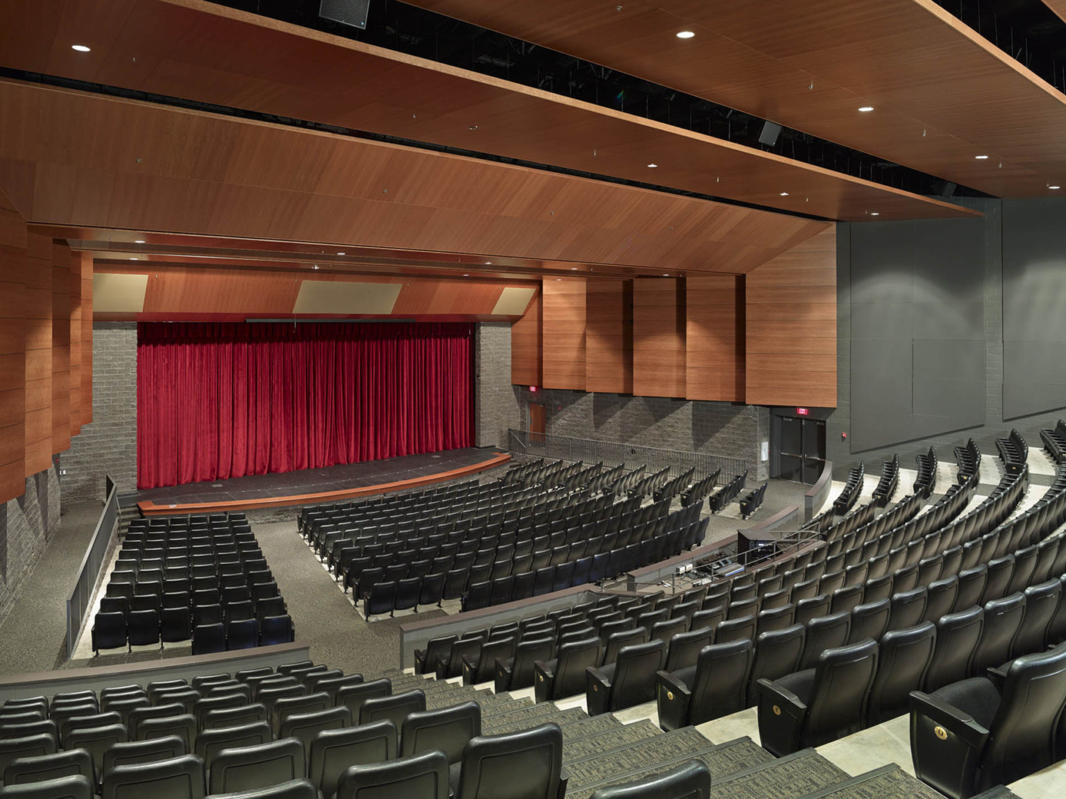 Auditorium with curved seating facing red curtained stage. Wood panels hang above audience and frame stage above stone wall
