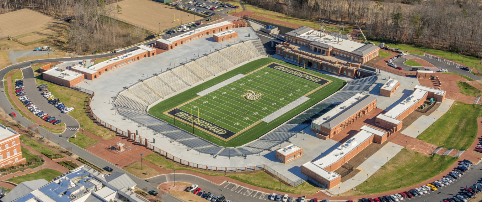 Aerial view of empty under grade level horseshoe shaped football stadium. Multistory brick building at far end of field