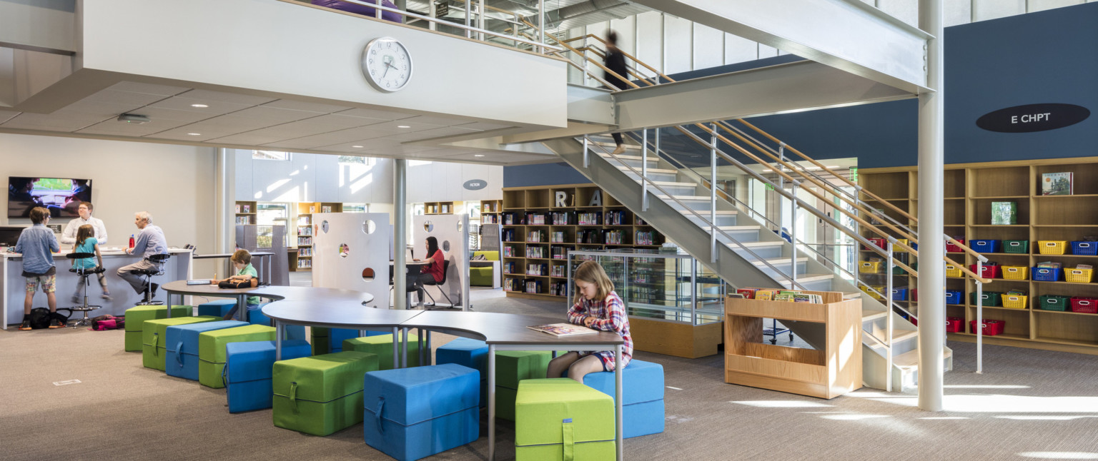 painted steel mezzanine with staircase over curved table with colorful soft seating next to bookshelf and clerestory windows