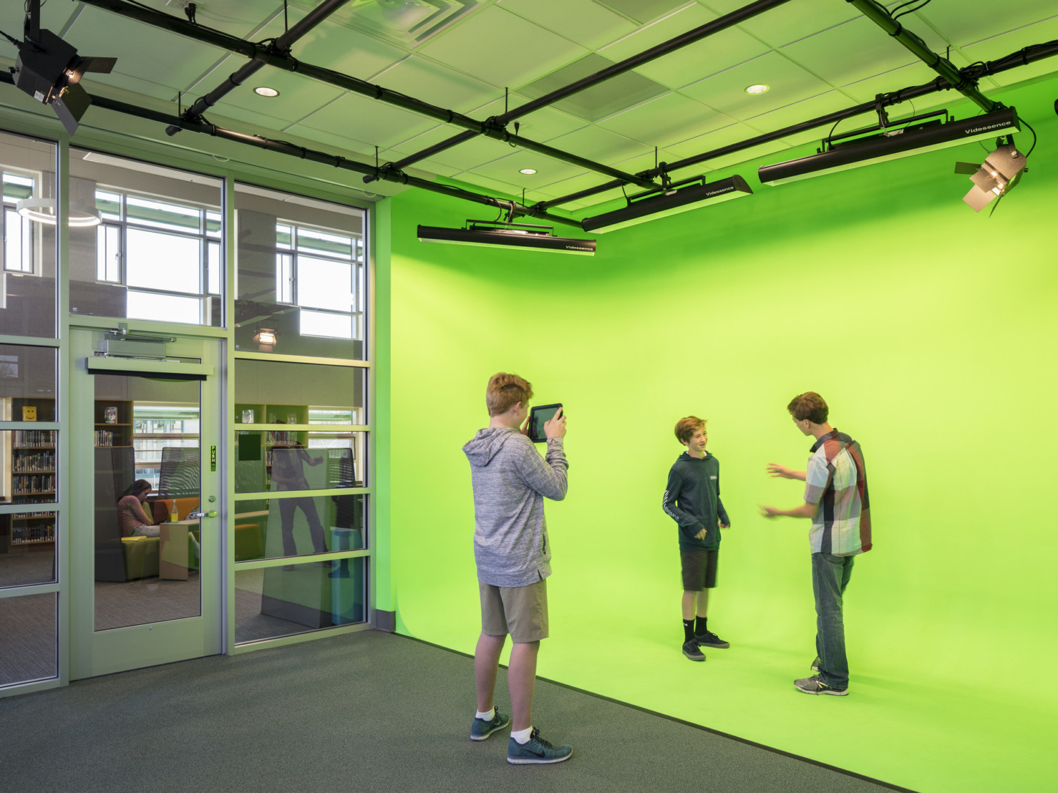 Green screen production set with open frame ceiling rigging and carpeted floor next to transparent glass wall system