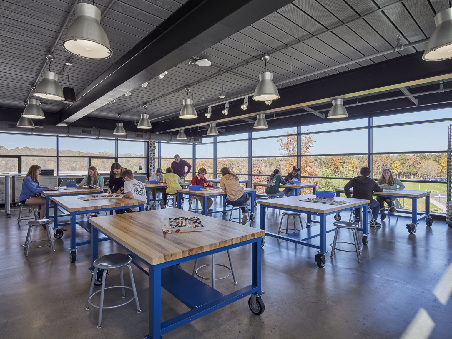 industrial tech classroom where students collaborate over movable tables with views to forest by floor to ceiling windows