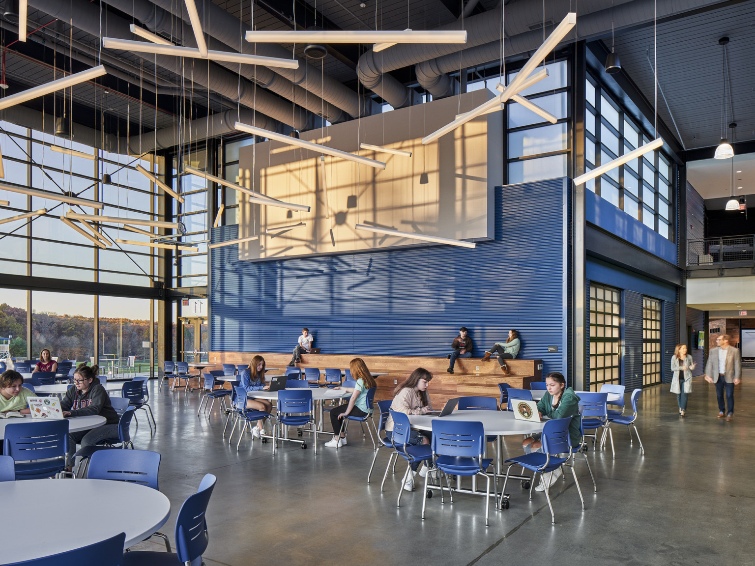 modern double height lunchroom with blue metal paneling, a wood learning stair, and floor to ceiling windows overlooking the football field