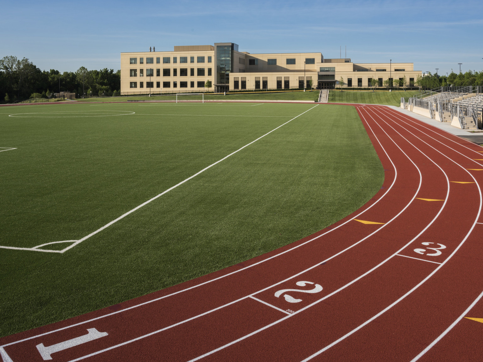 red colored track of a school's track and field athletic field, a light precast concrete building is in the distance
