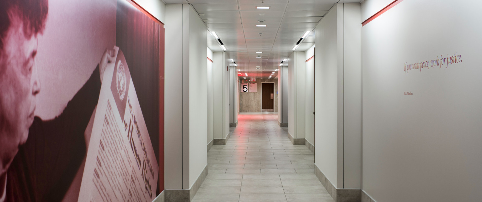 White hallway with red accent stripes. Front left section of hall has photo of a man with a piece of paper, opposite is quote