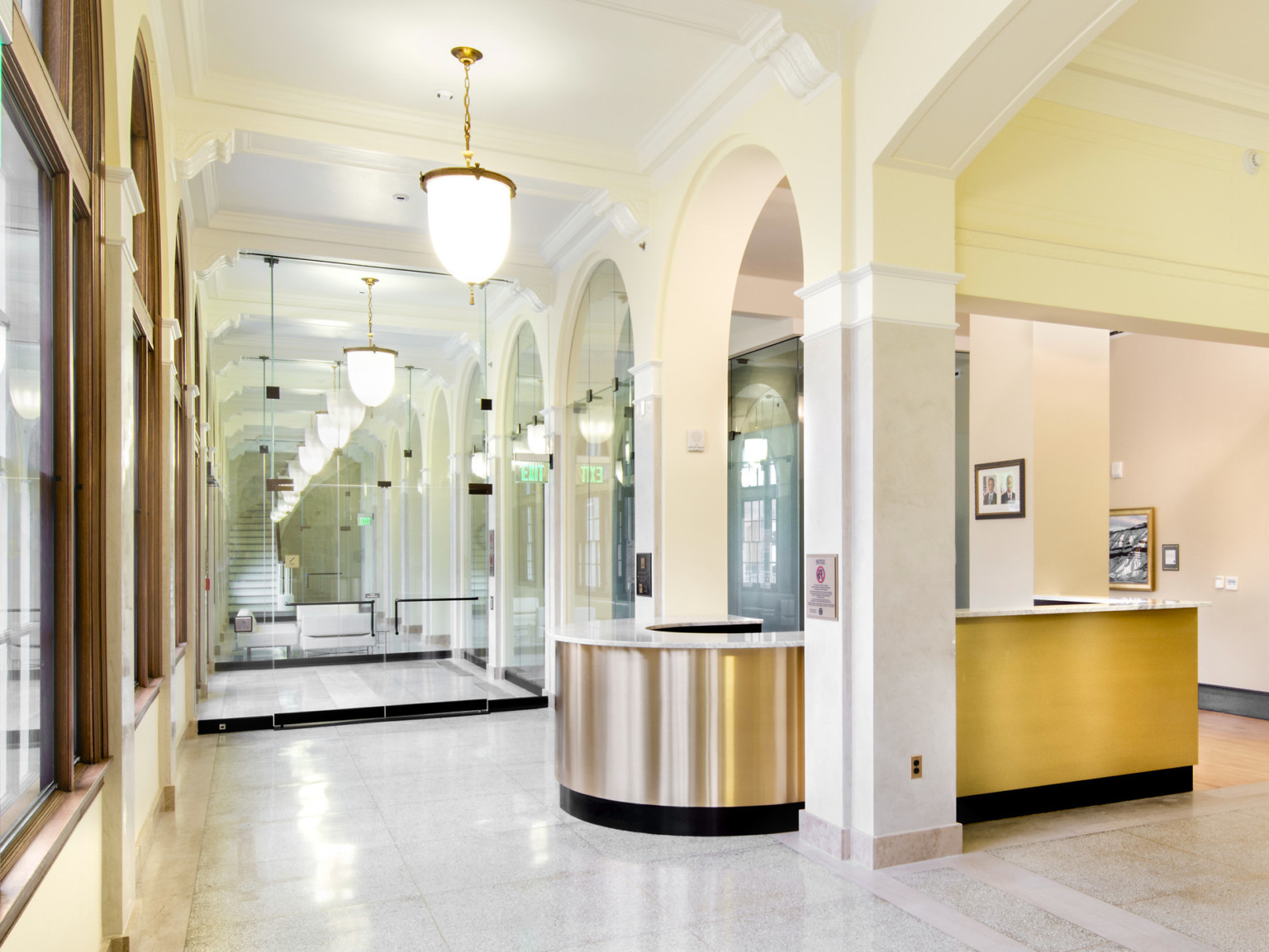 Gold reception desk with marble counter in front of a mirrored hallway with arched windows and hanging light