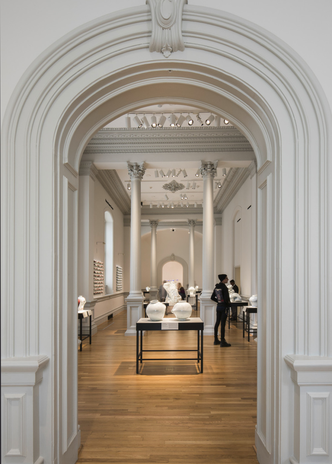 white art gallery with tall pillars displaying pottery on dark tables, framed by crown molding around doorway