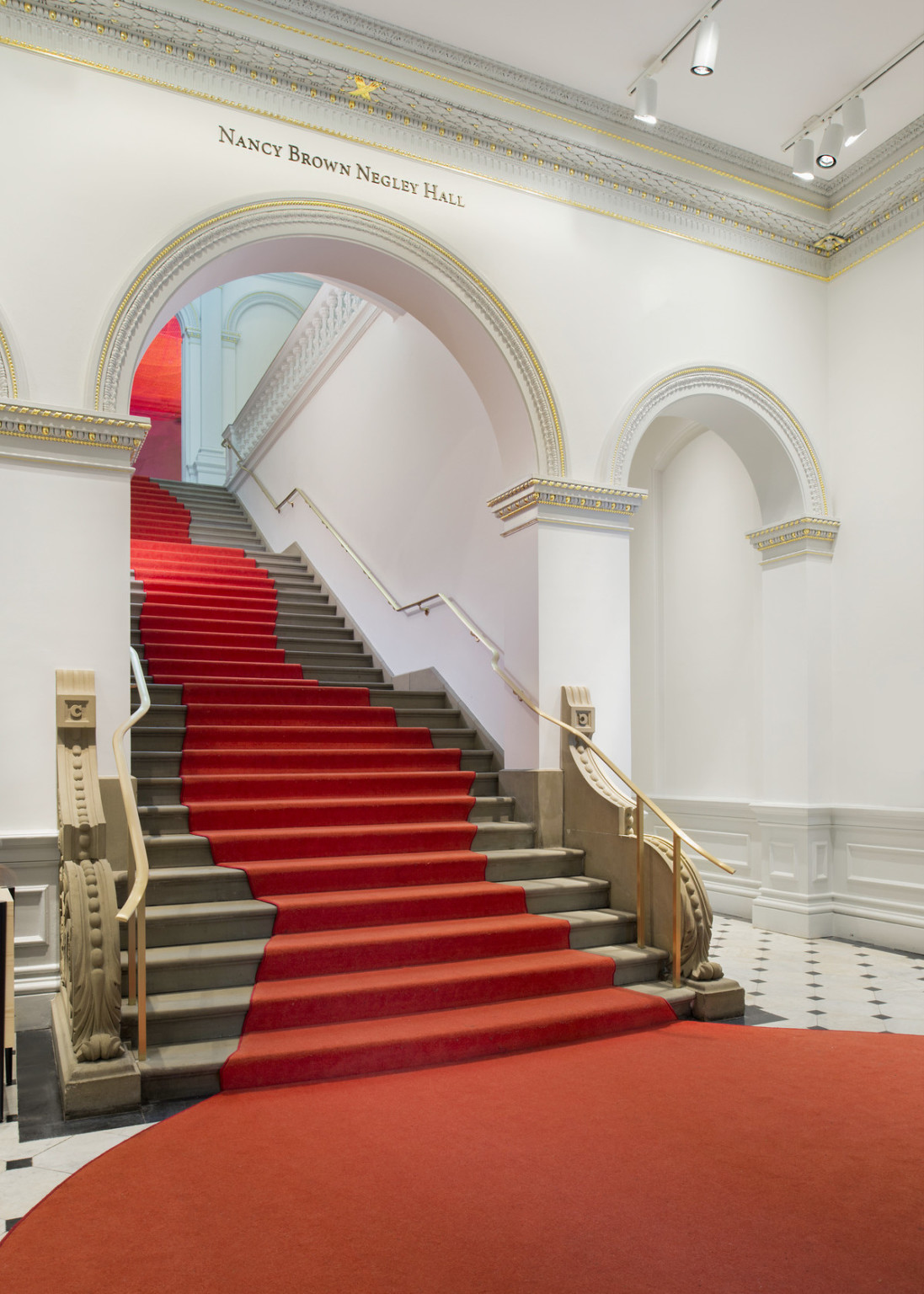 Grand staircase with curved red carpet that widens at base of stairs. Stairs pass through a white arch with gold molding